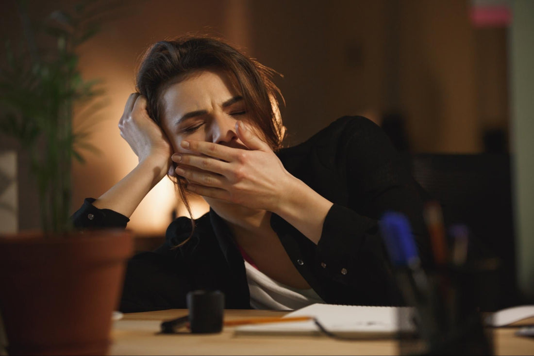 An exhausted woman yawning at her desk during a late-night work shift in a dimly lit office.